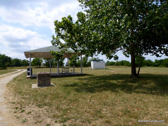 Prescott City Lake - Shelter - This is the picnic shelter at Prescott City Lake, with picnic tables, a trash can, and a stone hearth in the foreground, and the restrooms in the background. No water or electricity is available.